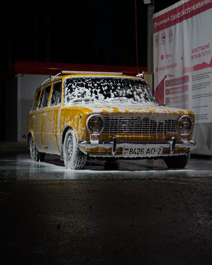 A classic yellow car covered in foam during a nighttime car wash. Vintage automotive style.